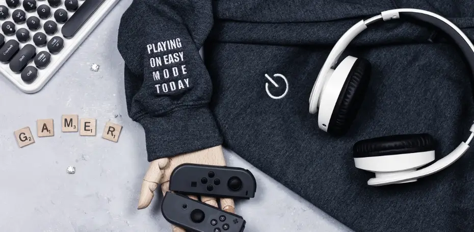 This image has a grey sweatshirt, switch controllers, white headphones, and a white keyboard with scrabble letters that spell gamer.